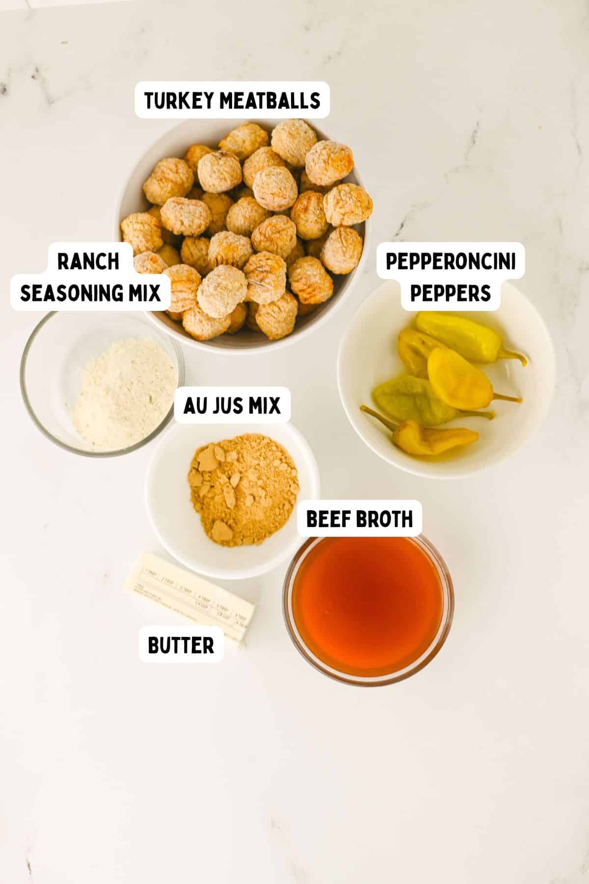 Five labeled ingredients on a white surface: turkey meatballs in a bowl, ranch seasoning mix in a small bowl, au jus mix in a bowl, beef broth in a bowl, a stick of butter, and pepperoncini peppers in a bowl.