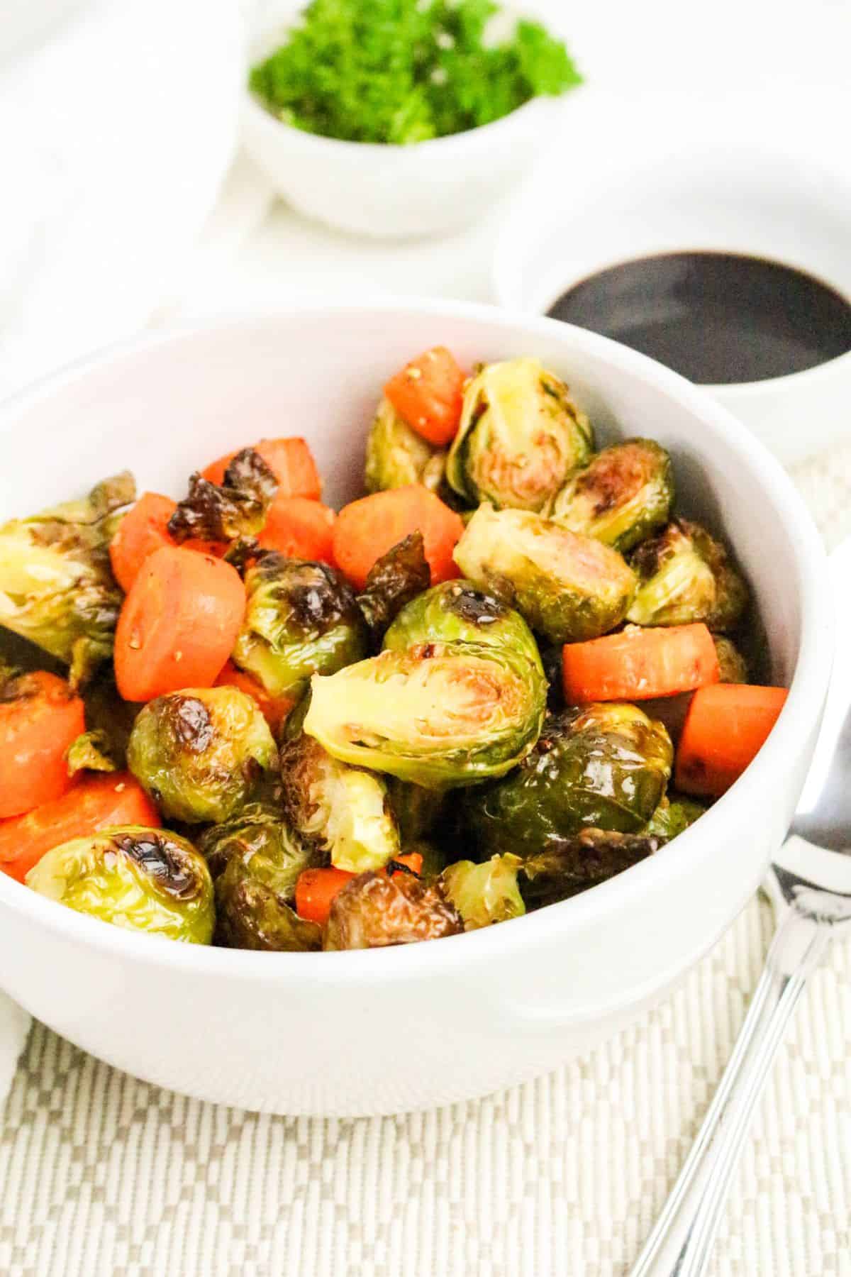 A white bowl filled with roasted Brussels sprouts and carrots sits on a light-colored placemat, with a spoon beside it. In the background, there is a small bowl of dark sauce and another bowl with fresh herbs.