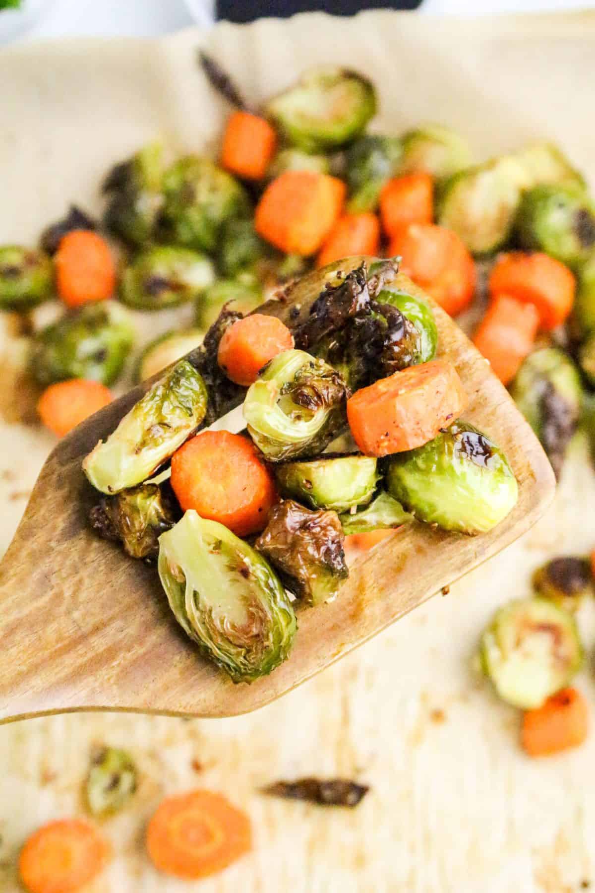 A wooden spoon holds roasted Brussels sprouts and carrot pieces above a baking sheet lined with parchment paper, with more roasted vegetables scattered in the background.