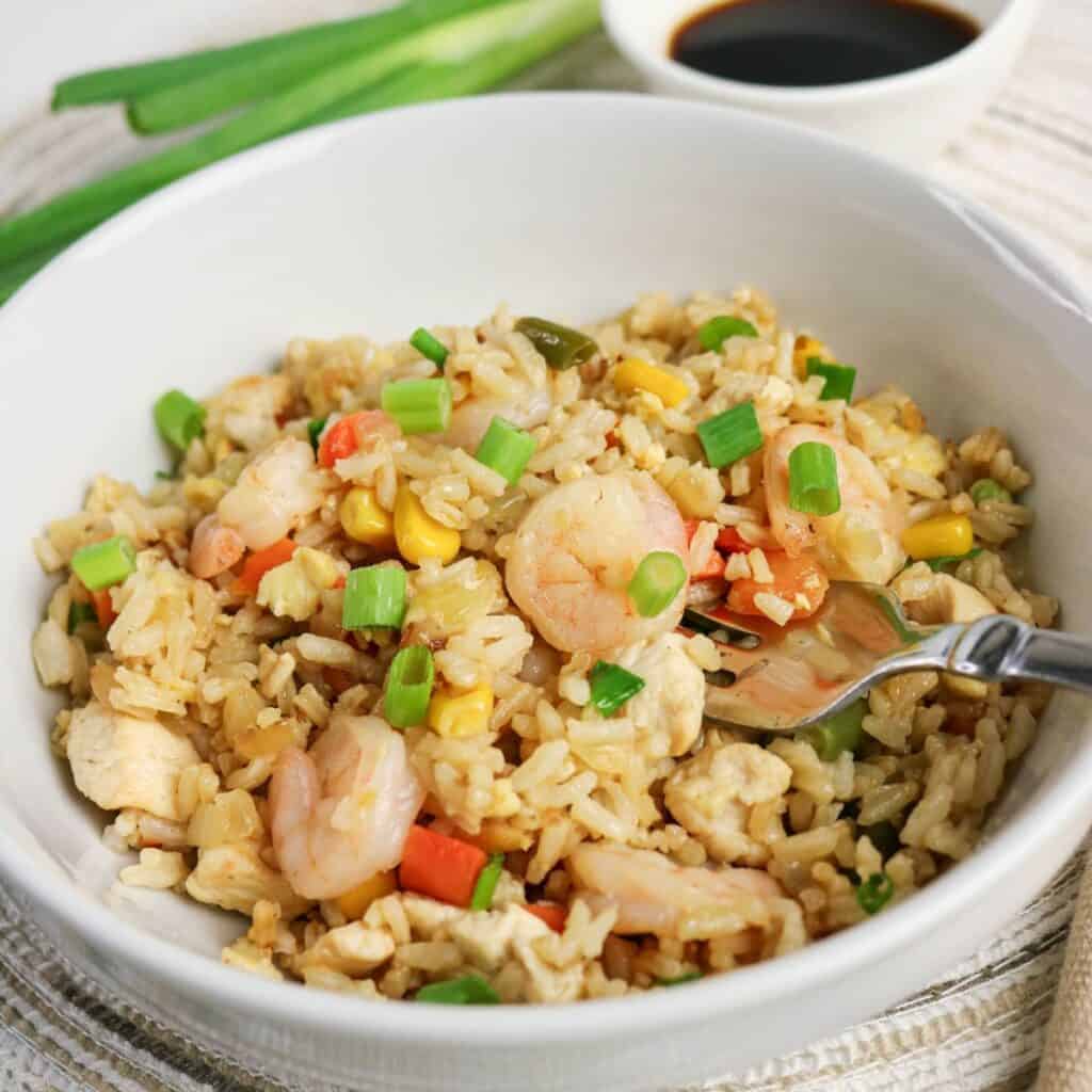 A white bowl filled with shrimp fried rice, including shrimp, rice, diced carrots, corn, green onions, and egg. A fork rests in the bowl. A cup of soy sauce and green onions are in the background.