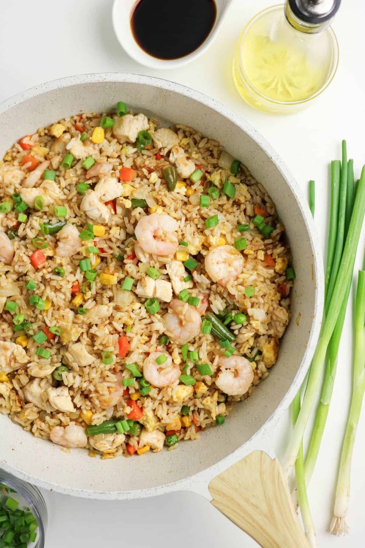 A skillet filled with shrimp fried rice containing shrimp, chicken, eggs, diced vegetables, and chopped green onions. Soy sauce, a bottle of oil, and fresh green onions are arranged nearby on a white surface.