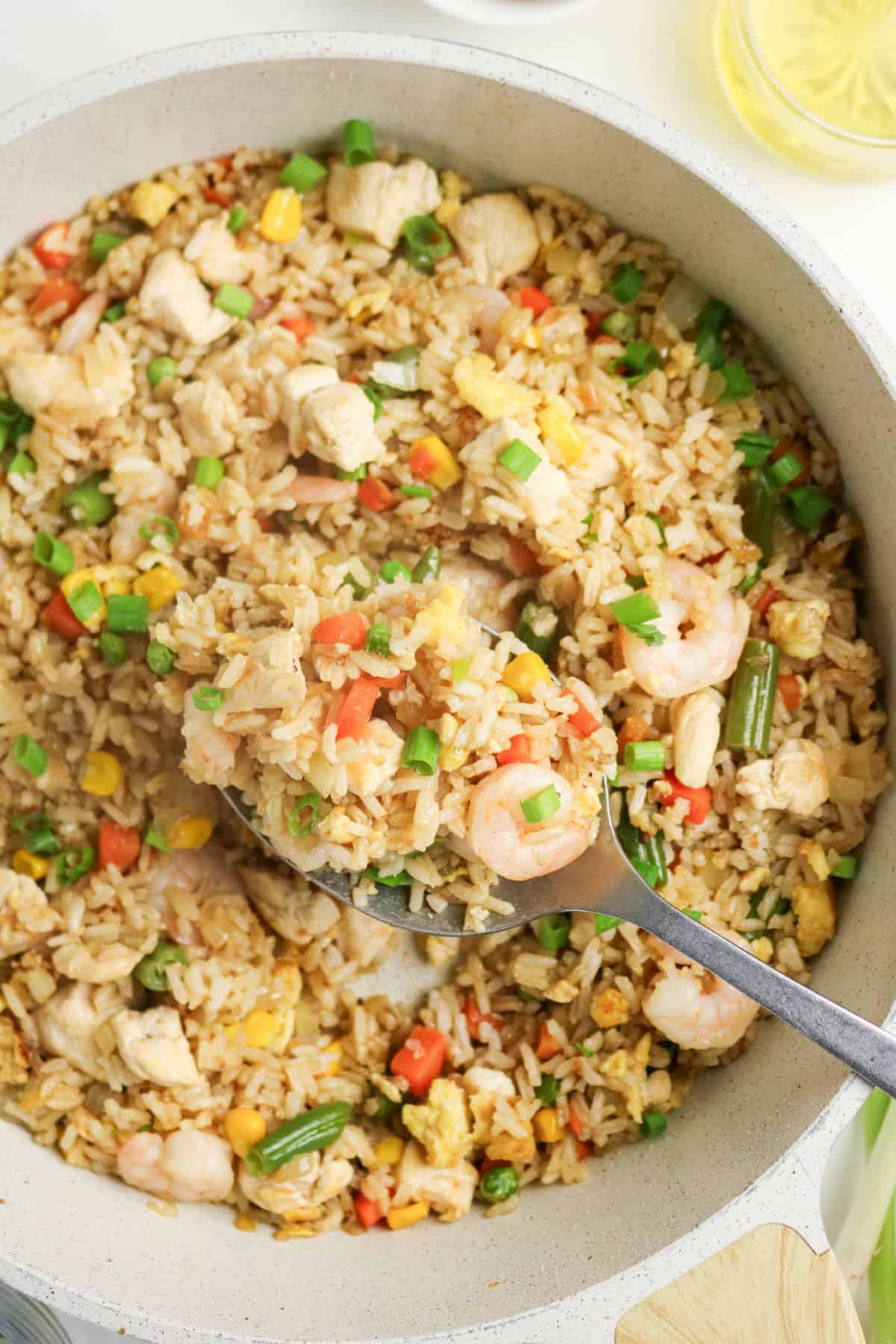 A pan filled with fried rice containing shrimp, chicken pieces, egg, green beans, corn, carrots, and green onions. A metal spoon is lifting a portion of the rice from the pan.
