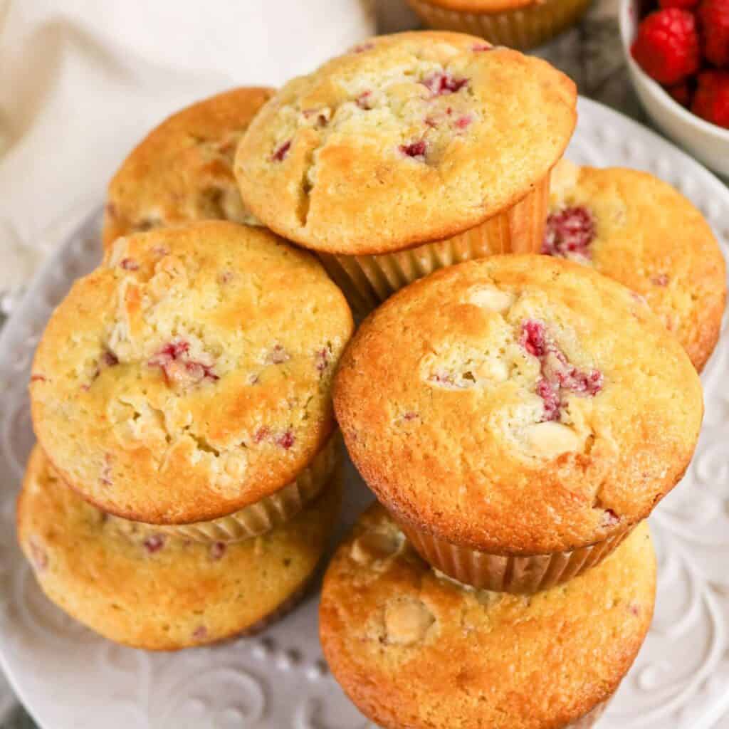 A plate stacked with several golden-brown muffins containing visible pieces of raspberries and white chocolate chunks, with a bowl of fresh raspberries in the background.