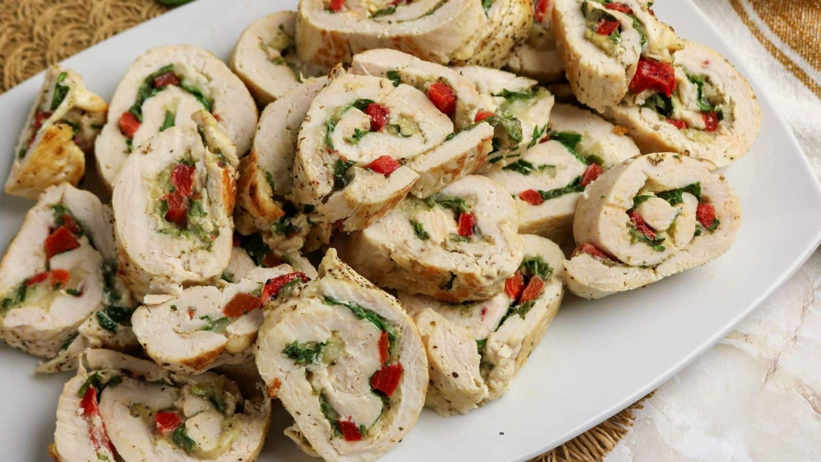 A white plate holds several pieces of rolled chicken breast stuffed with spinach, red bell peppers, and cheese, sliced into rounds and arranged in a pile.