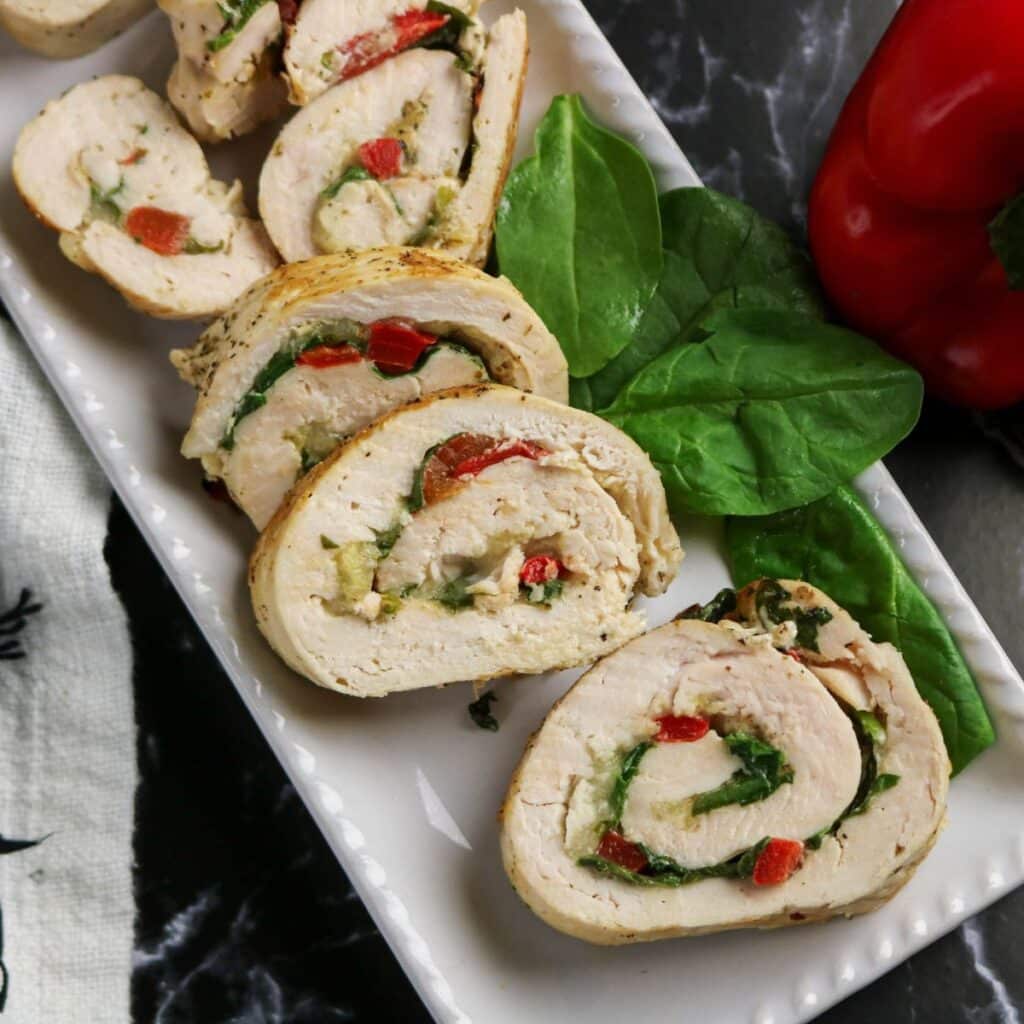 Sliced chicken rollatini stuffed with spinach and red bell pepper, arranged on a rectangular white plate with fresh spinach leaves beside the rolls. A red bell pepper is visible in the background.