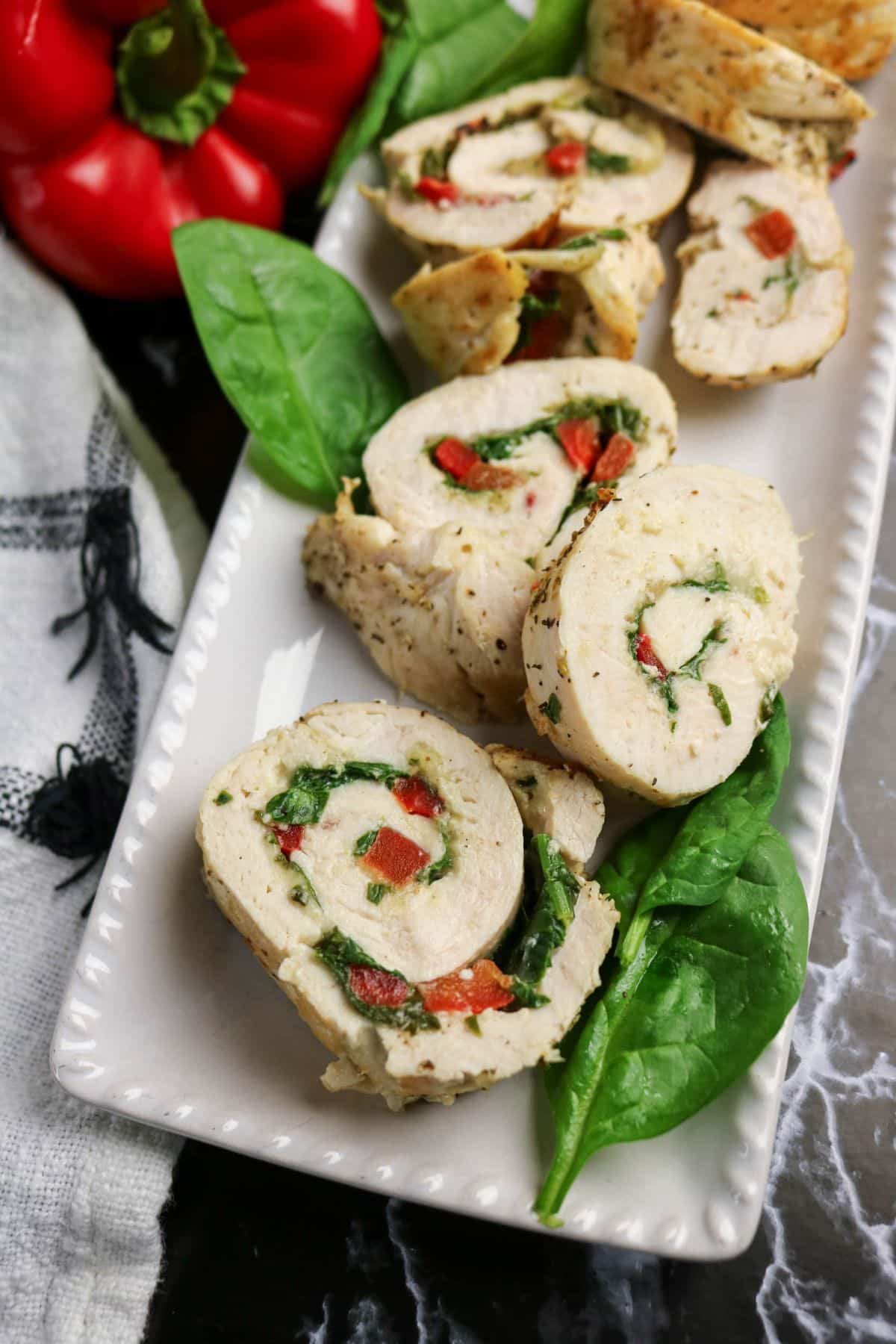 Sliced rolled chicken breast stuffed with spinach and red bell peppers is arranged on a white rectangular plate, with fresh spinach leaves and a red bell pepper beside the dish.
