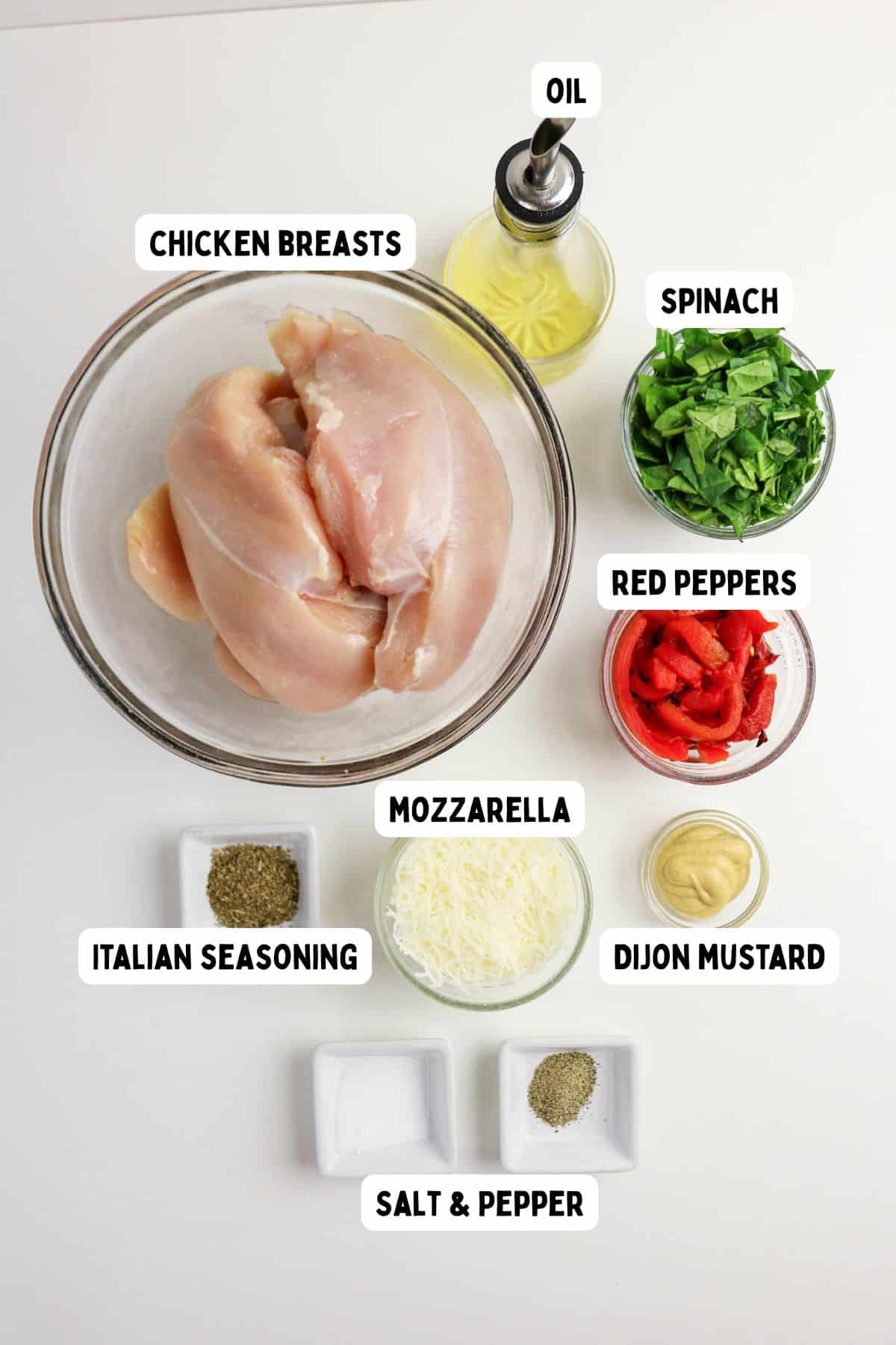 Overhead view of labeled ingredients on a white surface: raw chicken breasts in a bowl, oil, chopped spinach, sliced red peppers, shredded mozzarella, Italian seasoning, Dijon mustard, salt, and pepper.