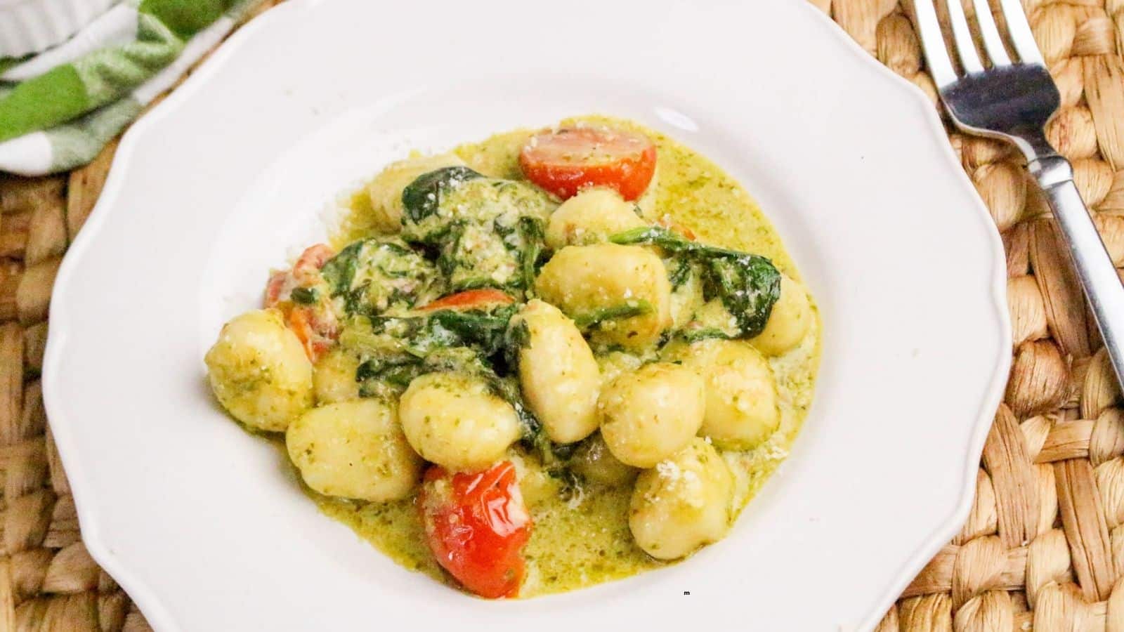 A white plate holds gnocchi in a creamy green sauce with spinach and halved cherry tomatoes. A fork is placed on a woven placemat next to the plate.