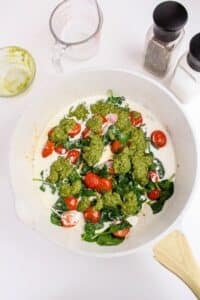 A white skillet containing cherry tomatoes, spinach, green pesto, and cream sits on a white surface. Nearby are a glass measuring cup, a green-stained bowl, and salt and pepper shakers.