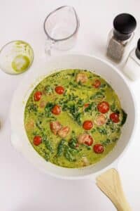 A white skillet with a creamy green sauce containing sliced sausage, cherry tomatoes, and spinach. Nearby are a glass measuring cup, an empty small bowl with green residue, and salt and pepper shakers.