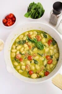 A white pot filled with gnocchi, halved cherry tomatoes, spinach, and a green sauce. Surrounding the pot are bowls of cherry tomatoes, spinach, grated cheese, and a pepper grinder on a white surface.