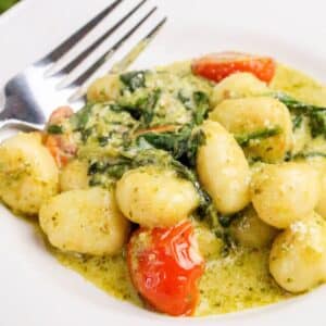 A plate of gnocchi in a creamy green pesto sauce with wilted spinach and cherry tomatoes, served next to a fork on a white plate.