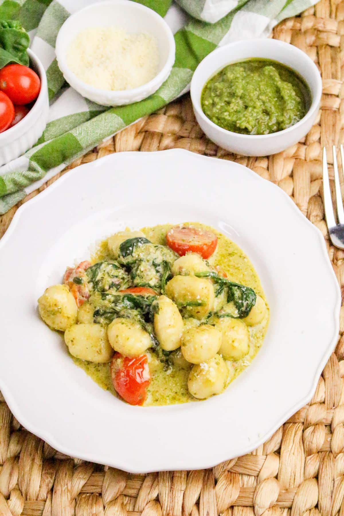 A white plate of gnocchi with cherry tomatoes, spinach, and pesto sauce sits on a woven placemat. Bowls of cherry tomatoes, grated cheese, and pesto are in the background. A fork is on the right side.