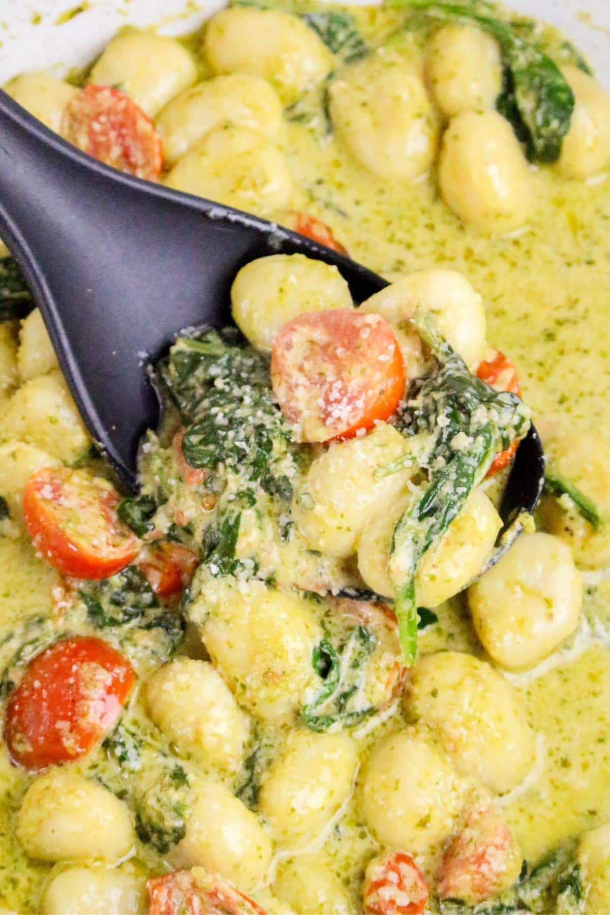 A close-up of gnocchi in a creamy green sauce with spinach and halved cherry tomatoes, being scooped with a black spoon. The dish is sprinkled with grated cheese.
