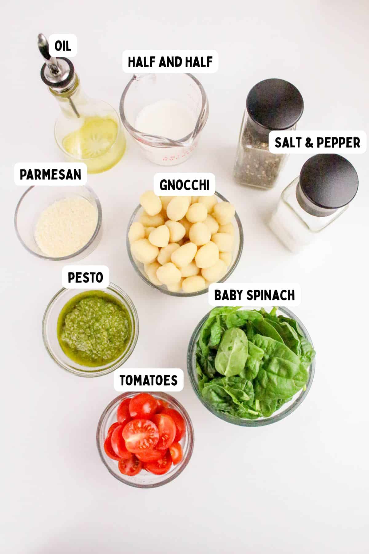 A top-down view of eight labeled bowls and containers with ingredients for a recipe: oil, half and half, salt and pepper, parmesan, gnocchi, pesto, baby spinach, and sliced tomatoes on a white surface.