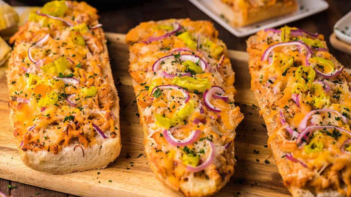 Three pieces of French bread pizza topped with melted cheese, sliced red onions, yellow peppers, herbs, and shredded chicken are arranged on a wooden cutting board. A plate with more pizza is in the background.