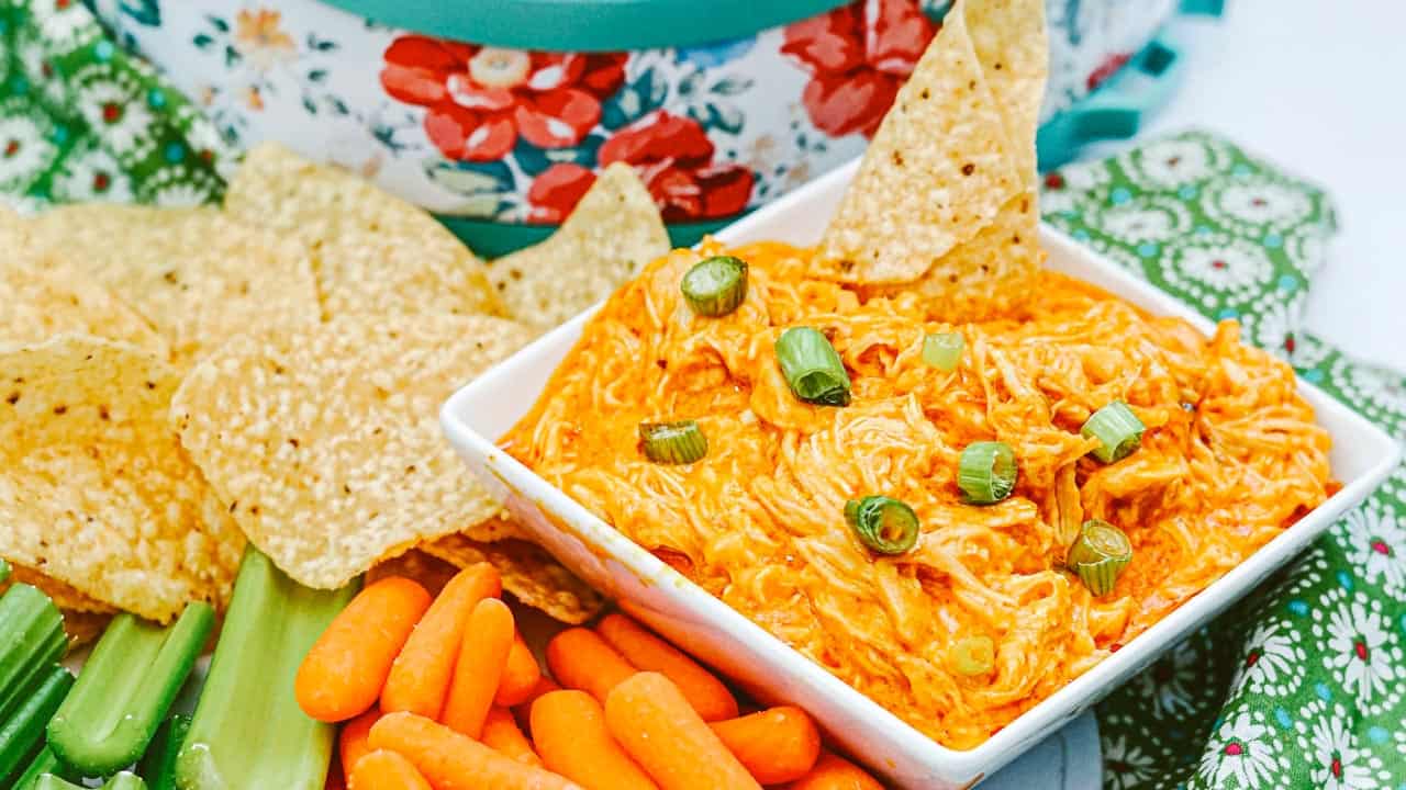 A square white bowl filled with creamy buffalo chicken dip topped with chopped green onions, surrounded by tortilla chips, baby carrots, and celery sticks on a green-patterned cloth.
