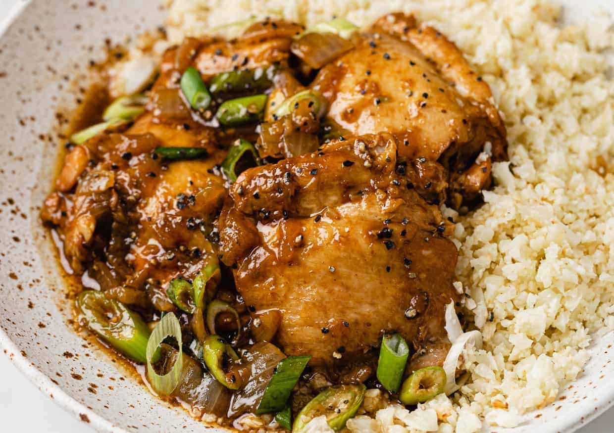 A close-up of browned chicken thighs topped with sliced green onions and a dark sauce, served next to a portion of cauliflower rice on a speckled white plate.
