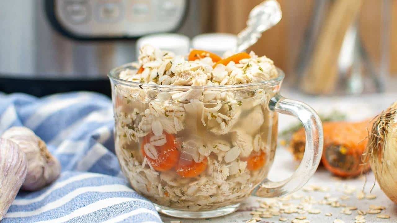 A clear glass mug filled with chicken soup containing shredded chicken, carrot slices, oats, and herbs, placed on a table with a blue and white striped cloth, garlic, onion, and a pressure cooker in the background.