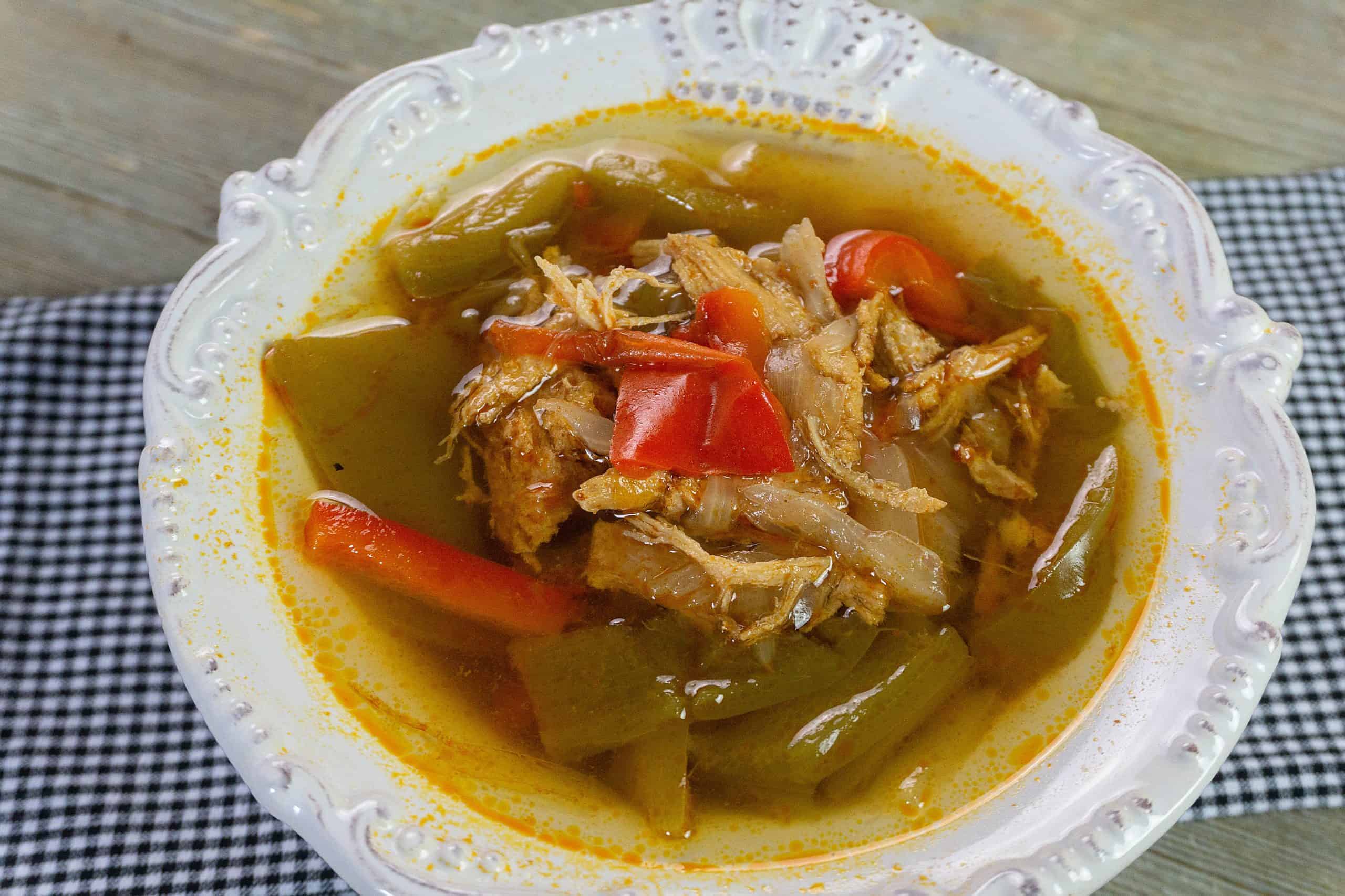 A white ornate bowl filled with fish soup containing chunks of fish, red and green bell peppers, and a yellowish broth, placed on a black and white checkered cloth on a wooden surface.