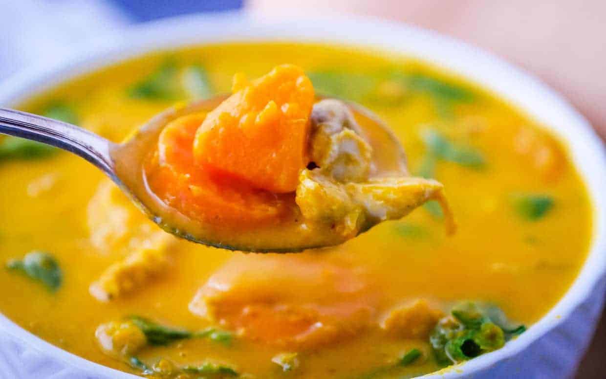 A close-up of a spoonful of yellow soup with chunks of orange sweet potato and green leafy vegetables, held above a bowl filled with the same soup.