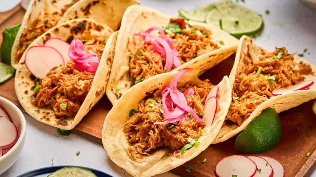 Four soft tacos filled with shredded meat, topped with pickled red onions and sliced radishes, are arranged on a wooden board with lime wedges and garnished with chopped herbs.