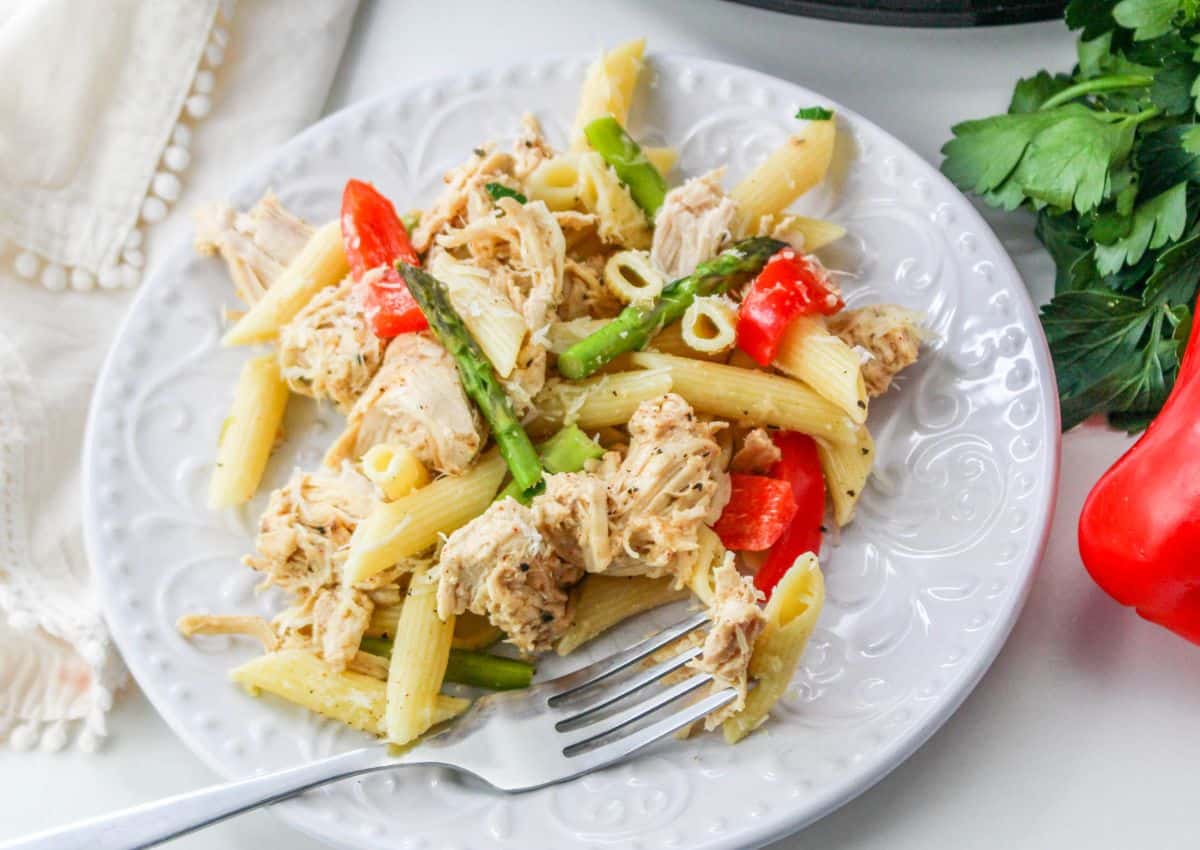 A white plate holds penne pasta mixed with shredded chicken, red bell peppers, and asparagus. A fork rests on the plate, and parsley and a red bell pepper are visible nearby.
