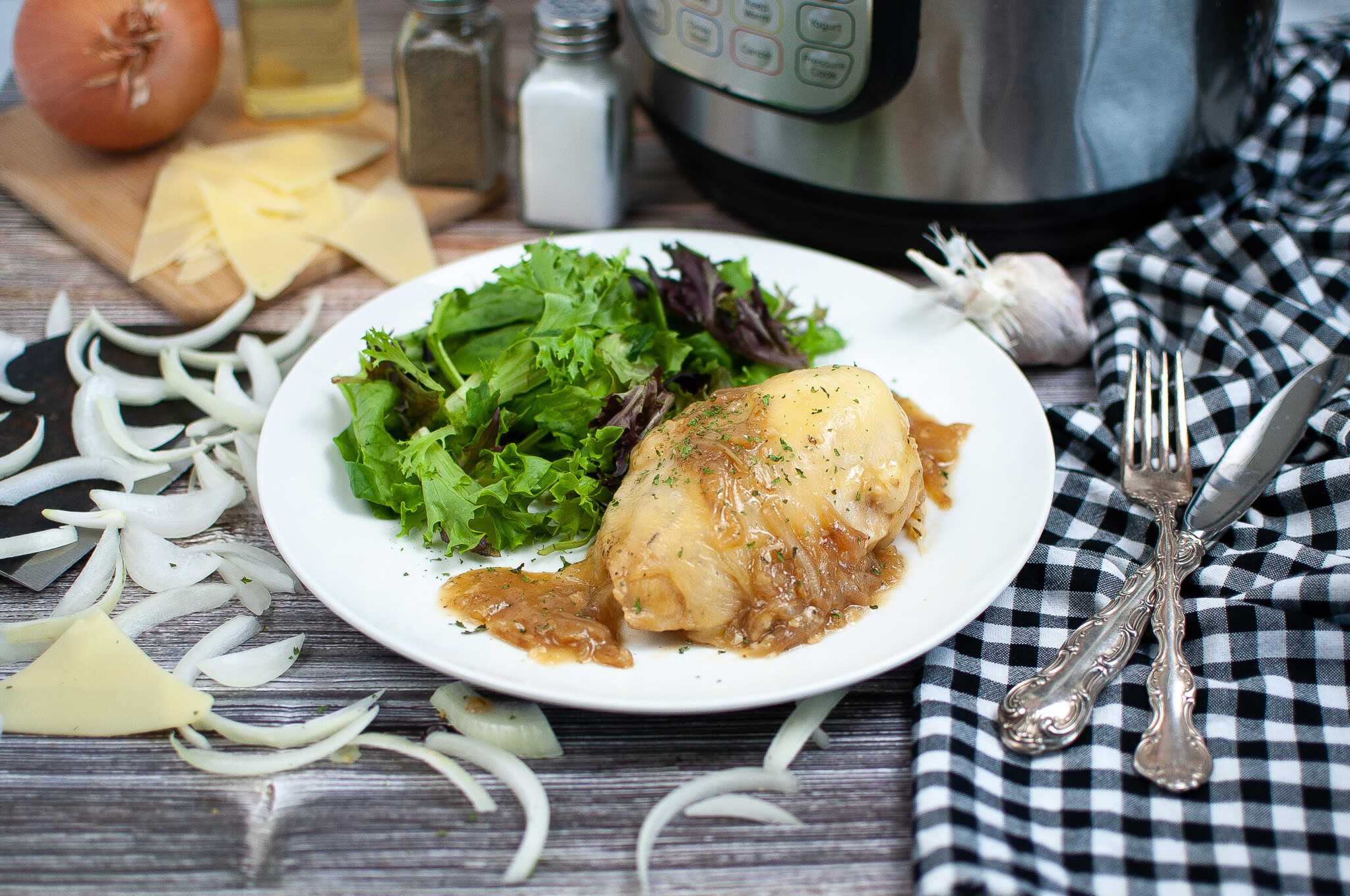 A white plate with baked chicken breast topped with sauce and a side of mixed green salad. Sliced onions and cheese are nearby, with utensils, a black-and-white checked napkin, and a pressure cooker in the background.