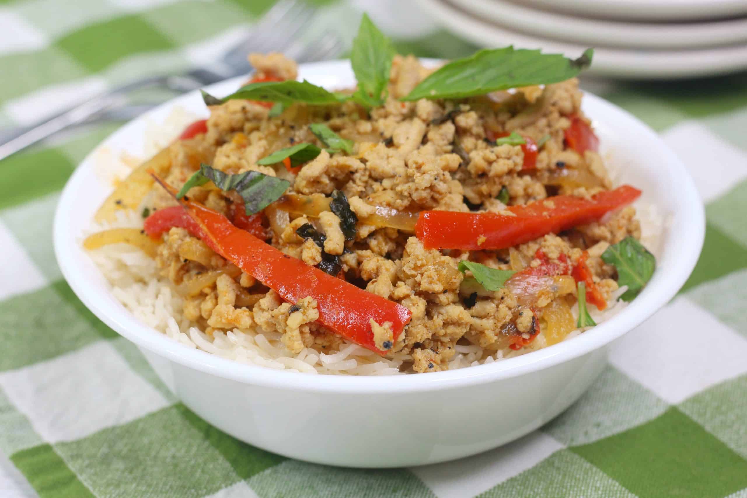 A white bowl filled with rice, topped with cooked ground meat, sliced red bell peppers, onions, and fresh herbs, sits on a green and white checkered tablecloth.