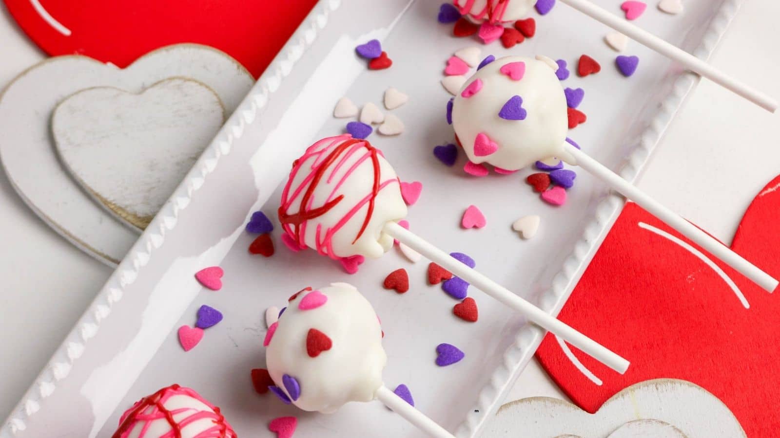 A rectangular white plate holds cake pops coated in white icing and decorated with red, pink, and purple heart-shaped sprinkles. The plate sits on a surface with heart-shaped decorations.