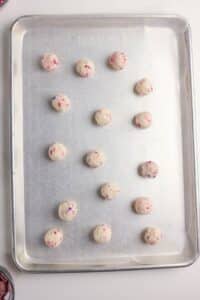 A metal baking sheet with fifteen evenly spaced balls of cookie dough containing colorful sprinkles, arranged in a grid pattern. The baking sheet is on a white surface.