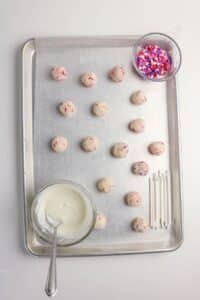 A baking sheet lined with parchment paper holds 16 unbaked sprinkle cake balls. Nearby are a bowl of white melted coating with a spoon, a bowl of colorful heart sprinkles, and a row of white lollipop sticks.
