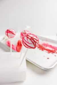 Three white cake pops on sticks with red and pink drizzle decoration. Two are standing in a white foam block, and one is in front. In the background, there is a tray with pink and red icing.