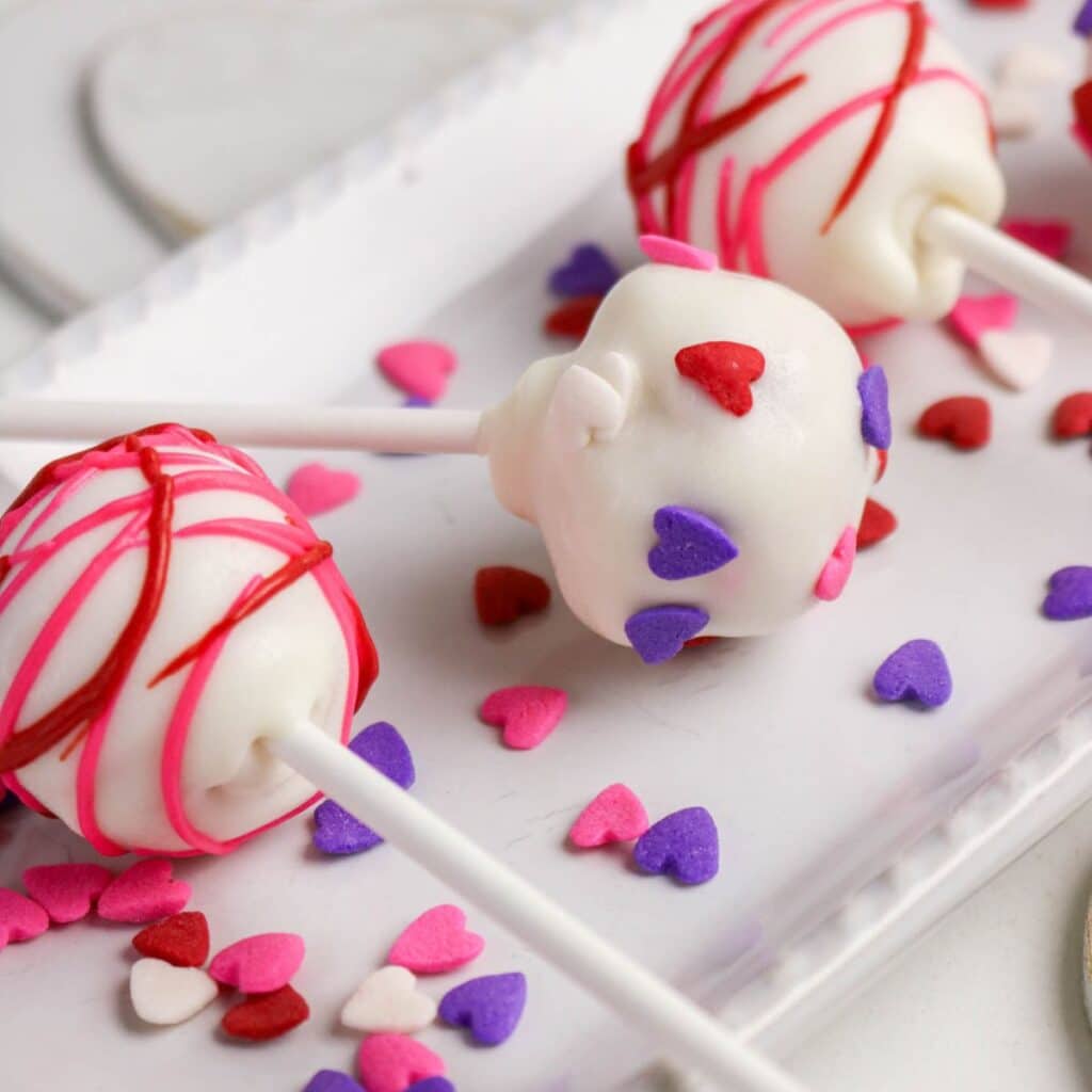Three cake pops coated in white chocolate with red and pink drizzle, decorated with pink, red, and purple heart-shaped sprinkles, are arranged on a white tray with additional heart sprinkles scattered around.