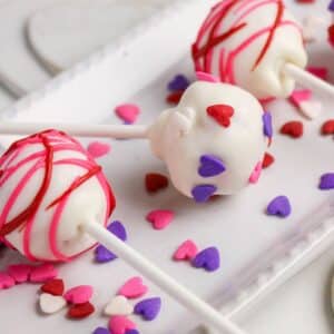 Three cake pops coated in white chocolate with red and pink drizzle, decorated with pink, red, and purple heart-shaped sprinkles, are arranged on a white tray with additional heart sprinkles scattered around.
