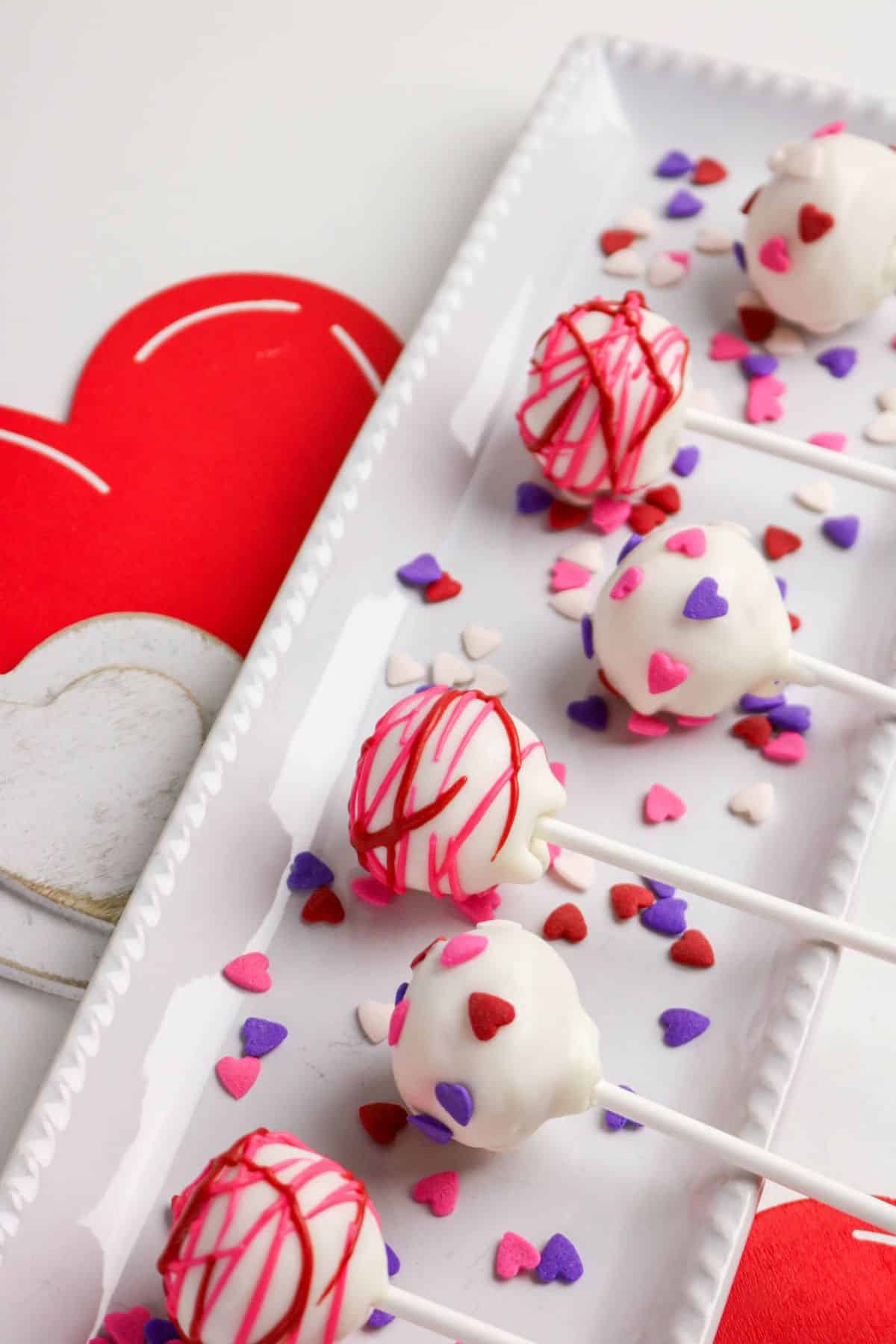 A rectangular white tray holds cake pops coated in white icing, decorated with red drizzle and pink, purple, and red heart sprinkles. Red and white heart-shaped cutouts are placed beside the tray.