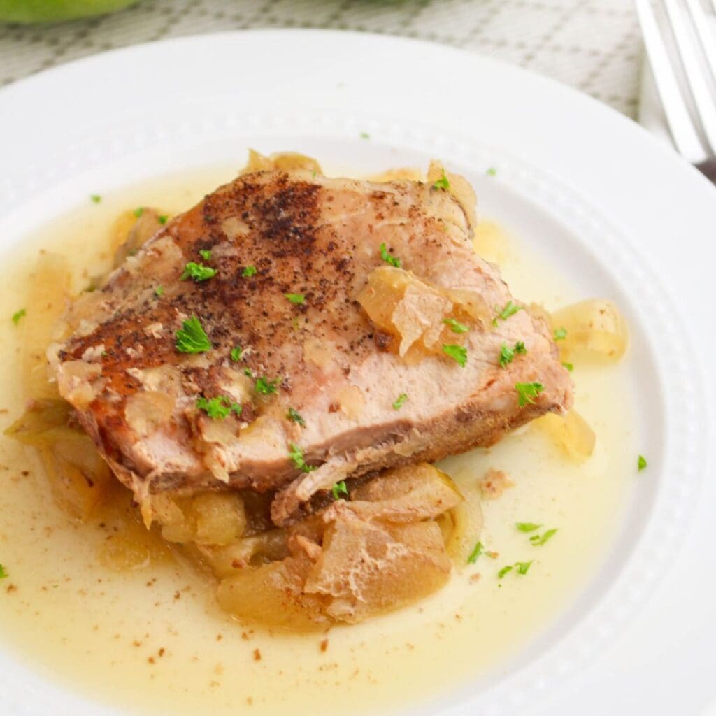 A pork chop topped with chopped parsley sits on a bed of cooked onions in a light, seasoned sauce on a white plate.