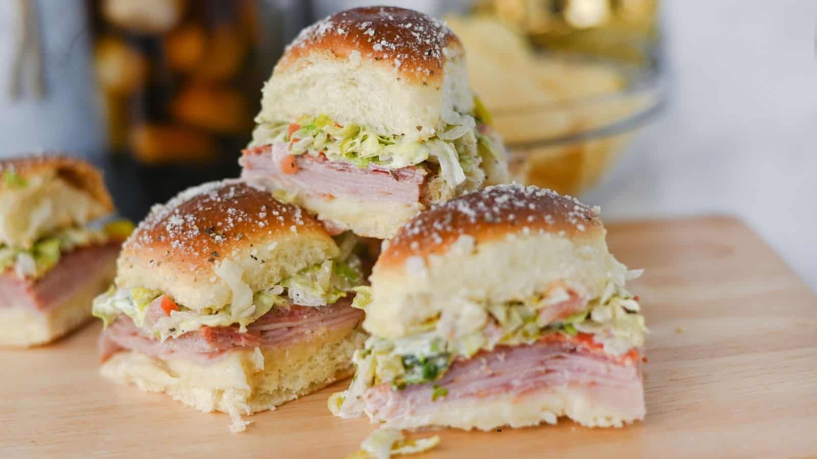 Three small deli meat sandwiches with lettuce and cheese are stacked on a wooden board. The sandwiches have soft, shiny buns topped with grated cheese or salt. A bowl of chips is visible in the background.
