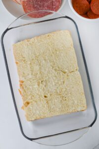 A glass baking dish containing six square pieces of plain sandwich bread. Slices of salami and pepperoni are visible in bowls nearby on a white surface.