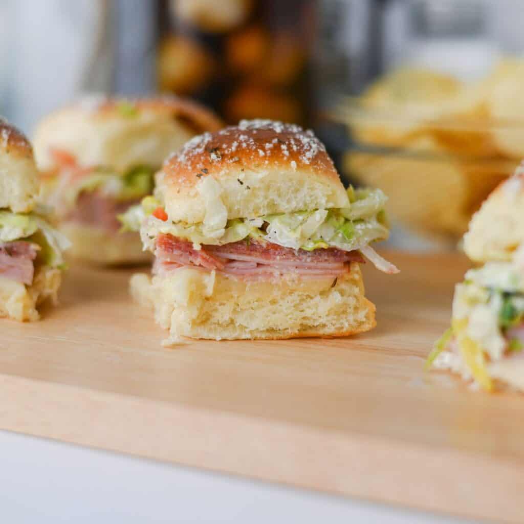 Three slider sandwiches with layers of deli meat, lettuce, and cheese are arranged on a wooden board. Potato chips are visible in the background.