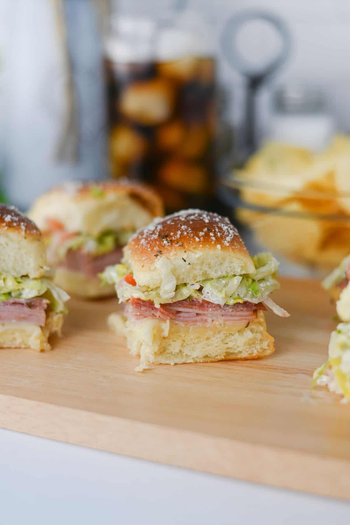A close-up of a sandwich slider with layers of meat, shredded lettuce, and cheese on a soft bun, placed on a wooden board. Other sliders and a glass of iced drink are blurred in the background.