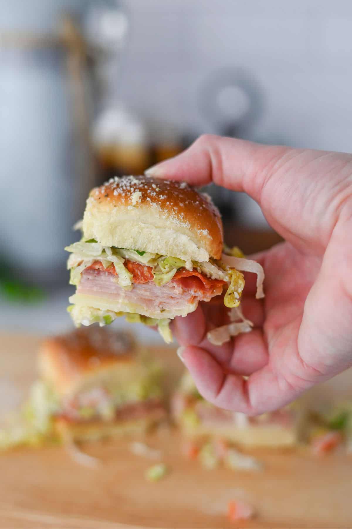 A hand holds a sandwich with layers of sliced deli meat, shredded lettuce, and a soft bun. Another sandwich and scattered lettuce pieces are visible on a wooden surface in the background.