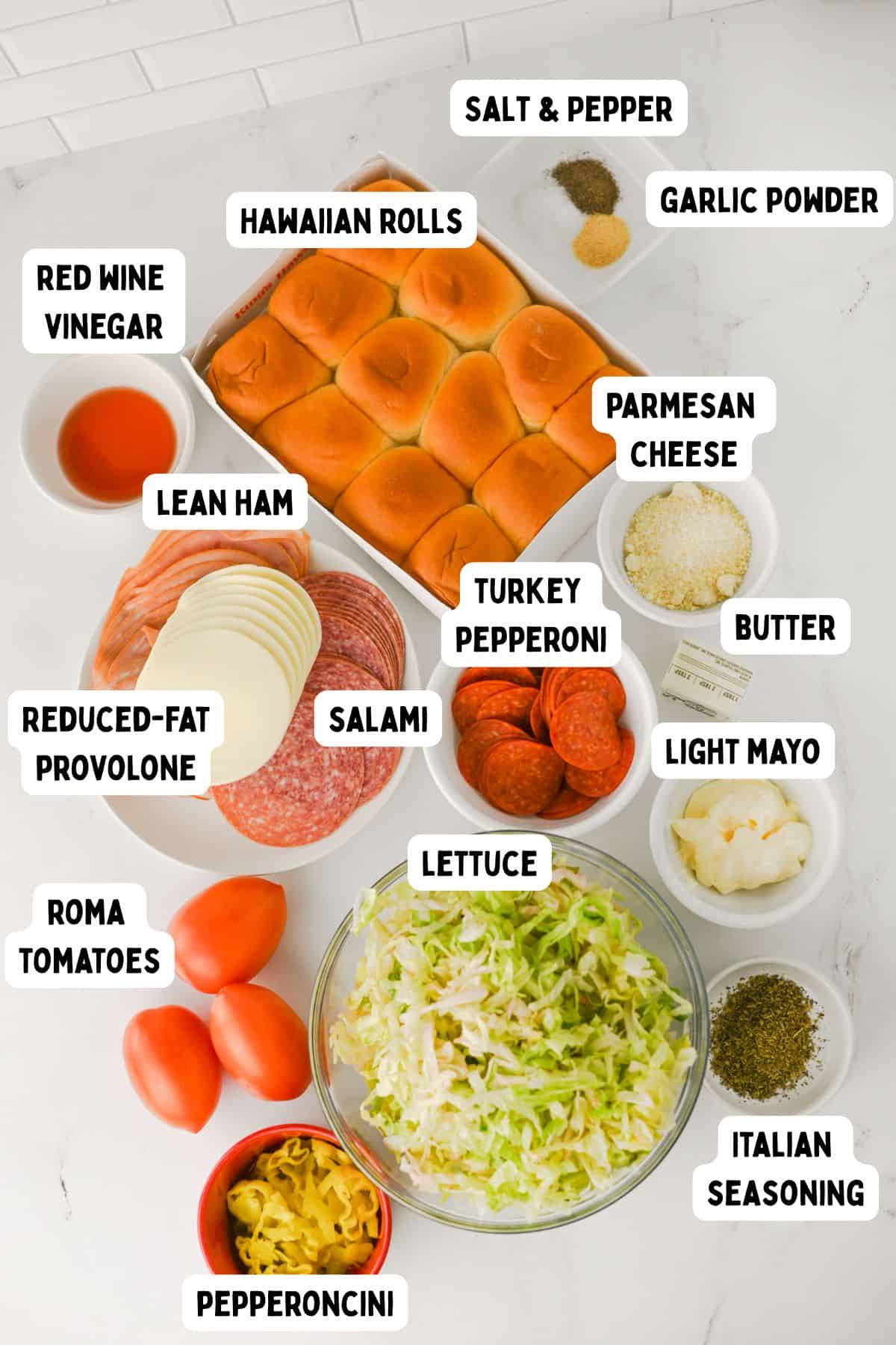 An overhead view of ingredients for sandwiches, including Hawaiian rolls, lean ham, salami, turkey pepperoni, provolone cheese, parmesan, butter, light mayo, lettuce, Roma tomatoes, pepperoncini, red wine vinegar, seasonings, and spices.