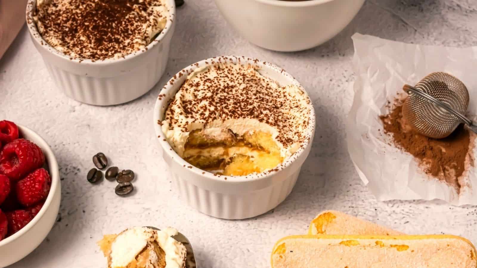 A ramekin of tiramisu with a spoonful removed sits on a light surface, surrounded by ladyfingers, loose cocoa powder with a sifter, fresh raspberries, coffee beans, and other bowls.