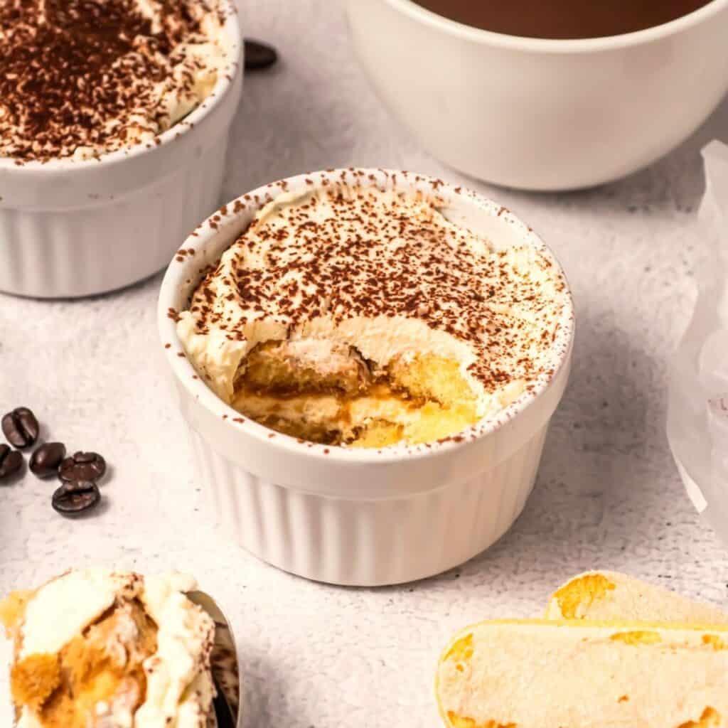 A white ramekin filled with tiramisu topped with cocoa powder. A spoonful has been taken out, revealing layers of cream and cake. Coffee beans, ladyfingers, and another ramekin are nearby on a light surface.