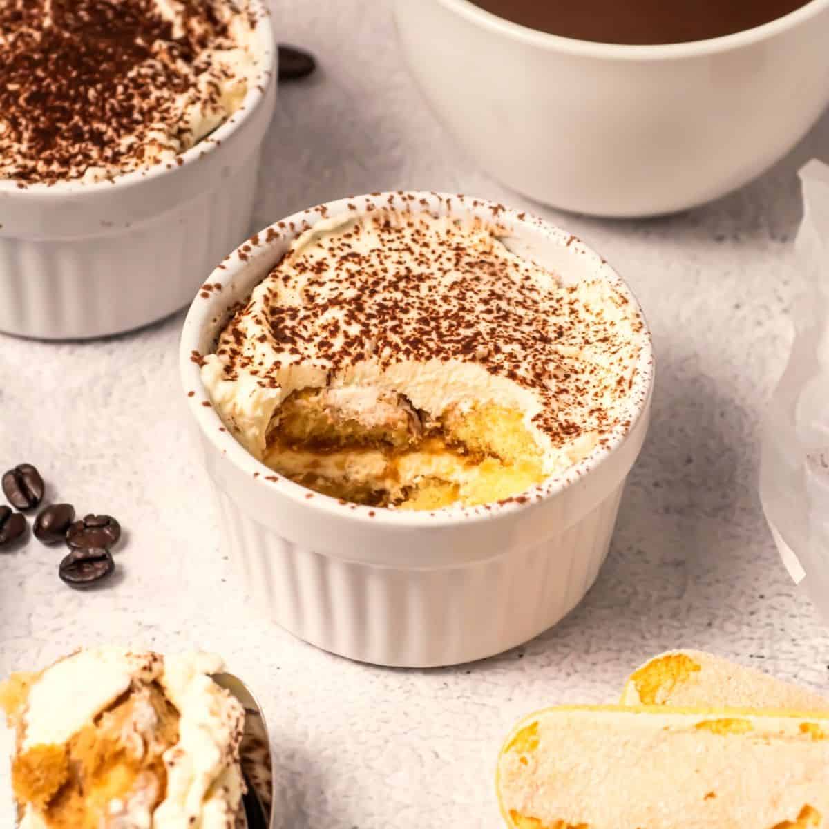 A white ramekin filled with tiramisu topped with cocoa powder. A spoonful has been taken out, revealing layers of cream and cake. Coffee beans, ladyfingers, and another ramekin are nearby on a light surface.