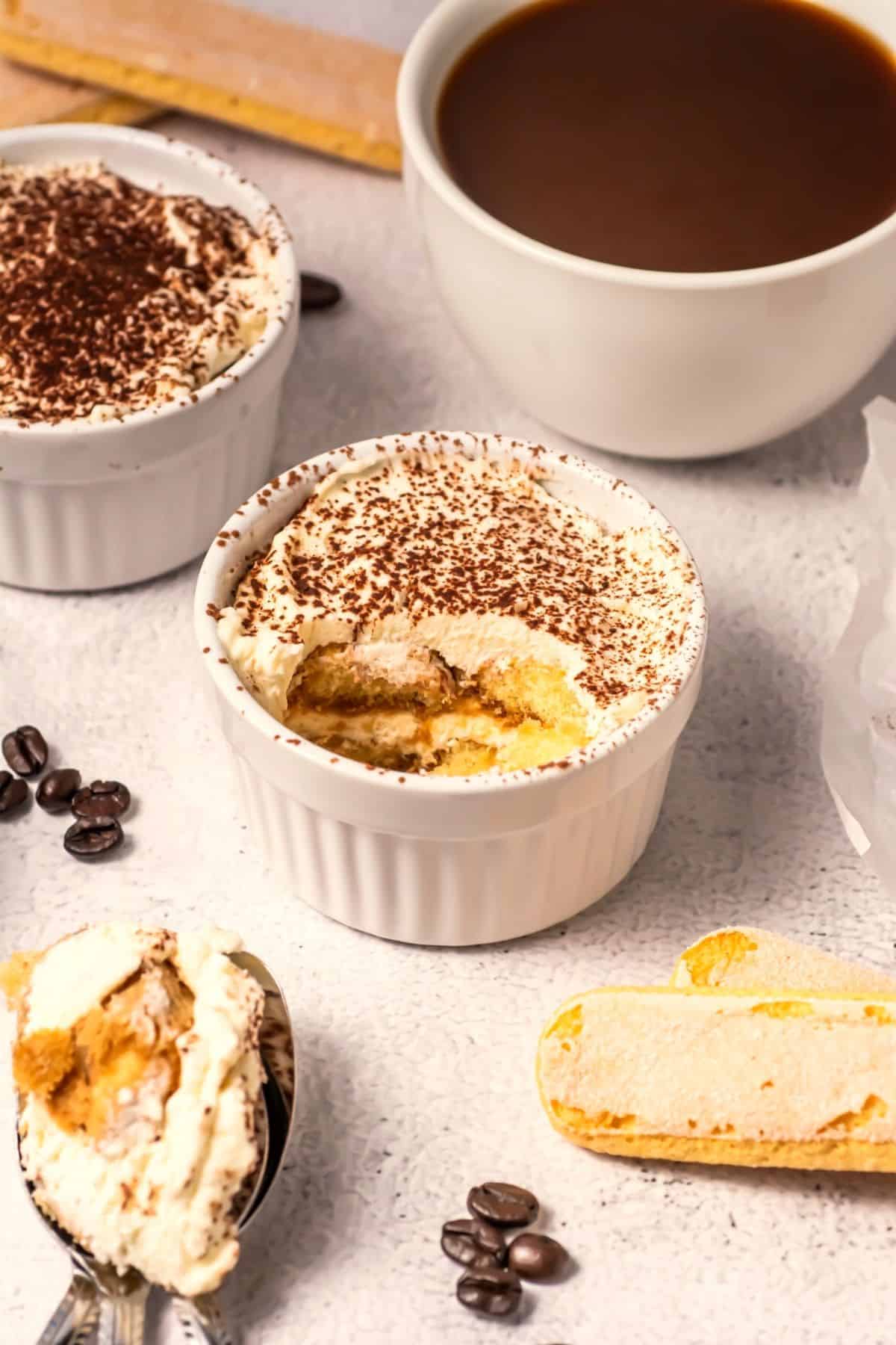 Two ramekins of tiramisu, one with a spoonful removed, sit on a light surface next to a cup of coffee, coffee beans, and ladyfinger biscuits. A spoon with tiramisu is in the foreground.
