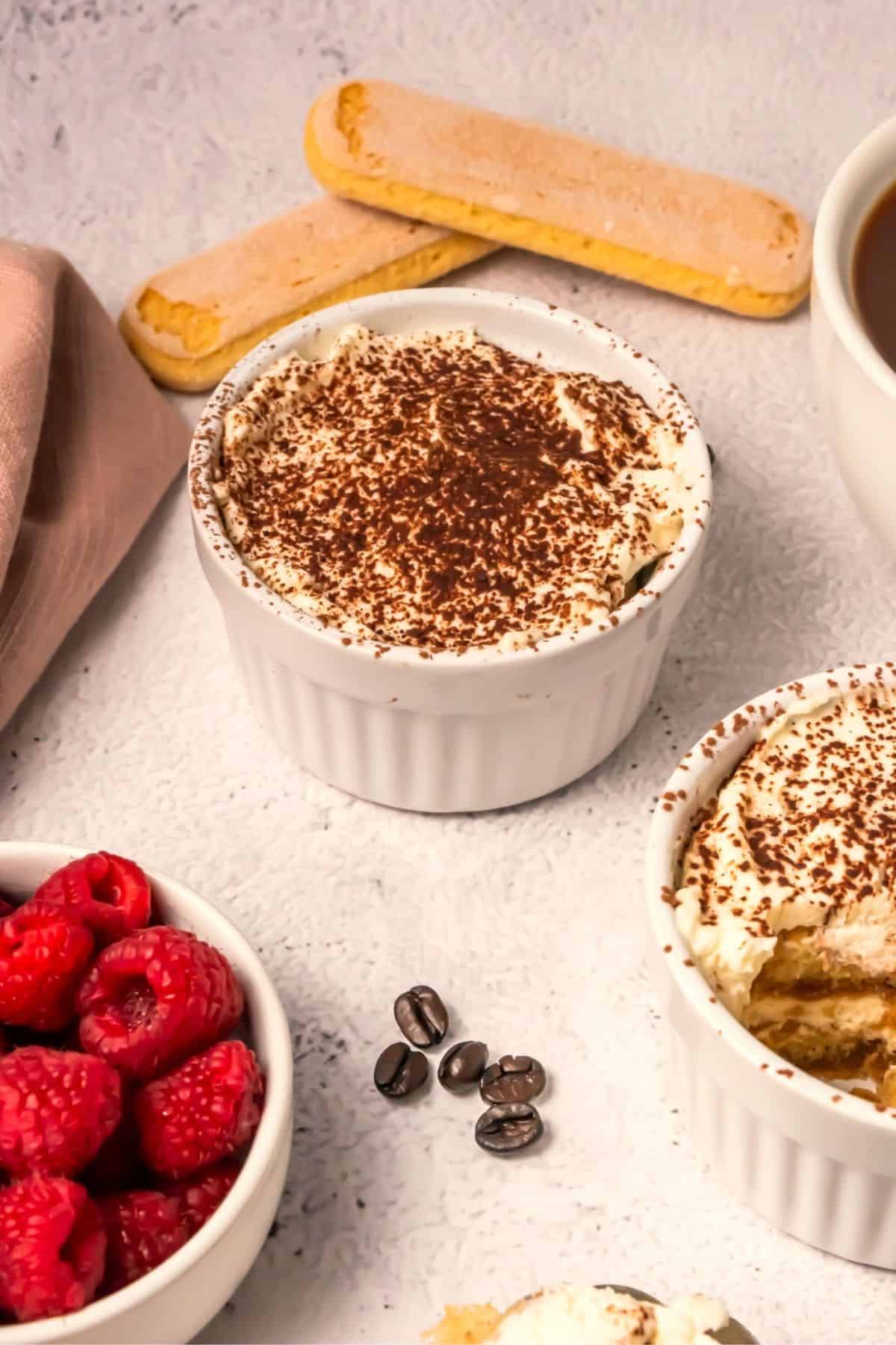 A white ramekin filled with tiramisu topped with cocoa powder sits on a light surface. Nearby are fresh raspberries, ladyfingers, coffee beans, and a cup of coffee.