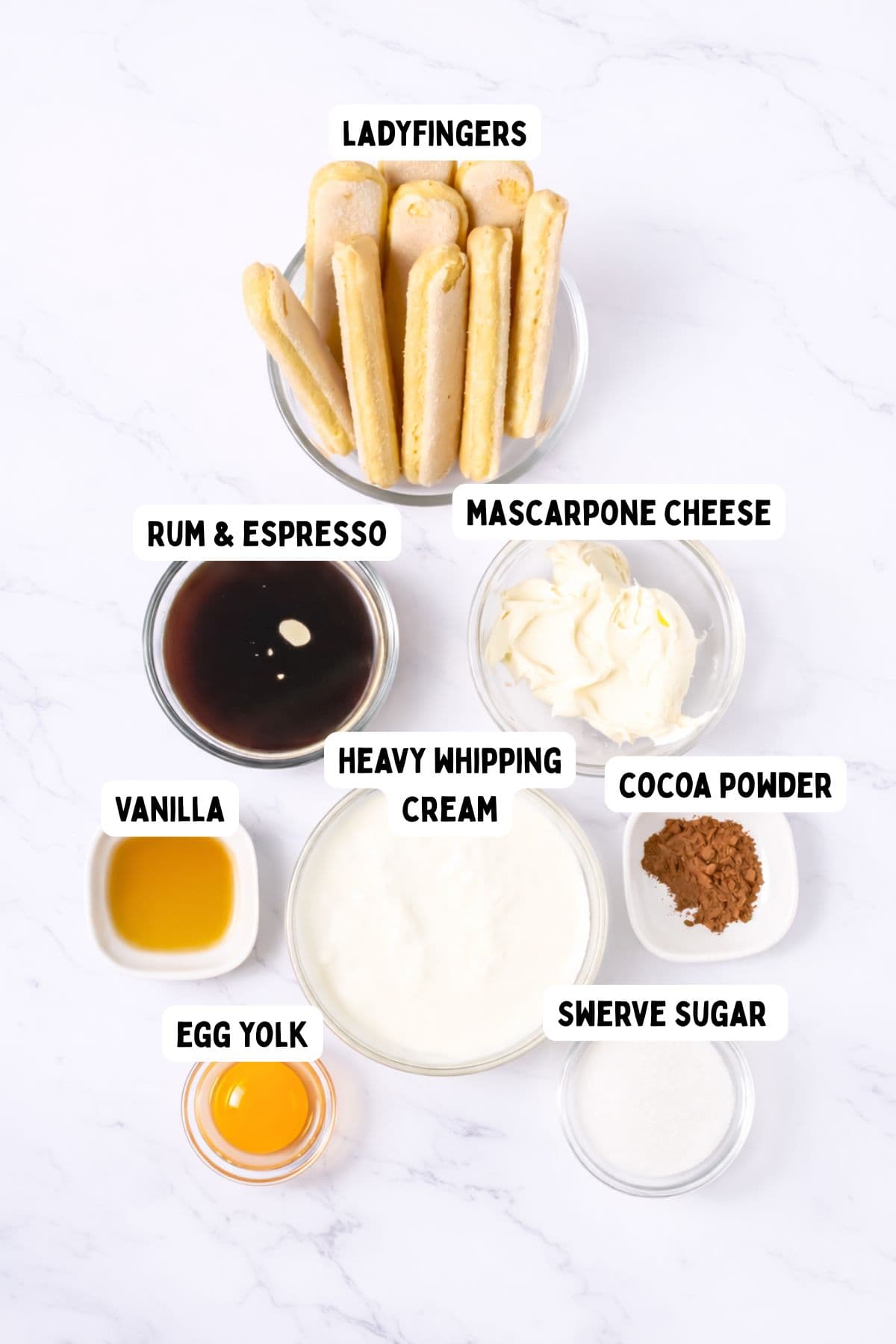 A top-down view of labeled ingredients for tiramisu, including ladyfingers, mascarpone cheese, rum and espresso, heavy whipping cream, cocoa powder, vanilla, egg yolk, and Swerve sugar, all in separate bowls on a white surface.
