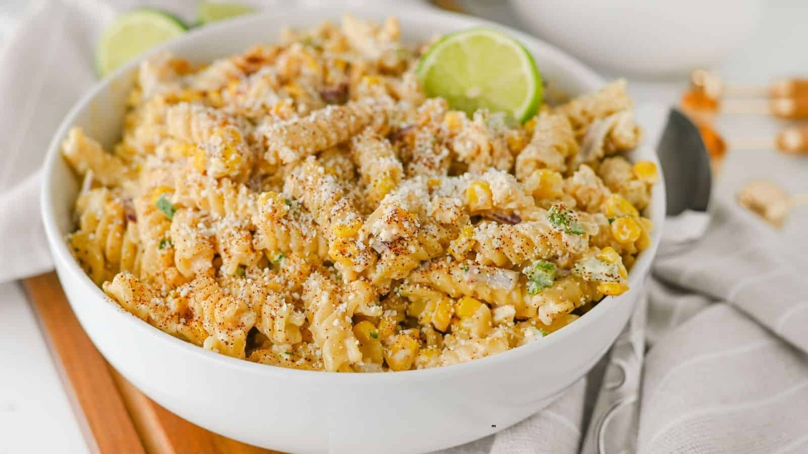 A white bowl filled with rotini pasta mixed with corn, green herbs, grated cheese, and spices, garnished with two lime slices, sits on a wooden board next to a gray-striped napkin and cutlery.
